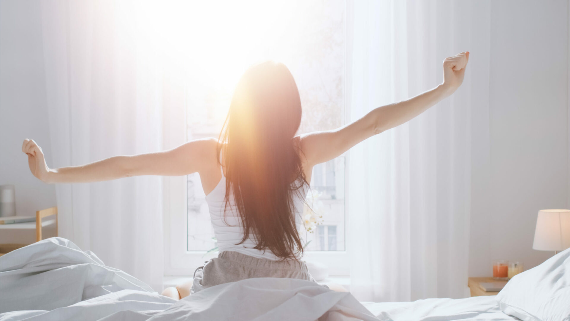 Young woman stretching sitting in bed, sun shining in her room
