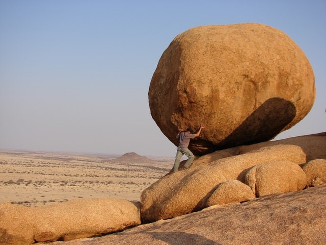 Mann in der Wueste, der versucht ein riesigen Stein zu halten