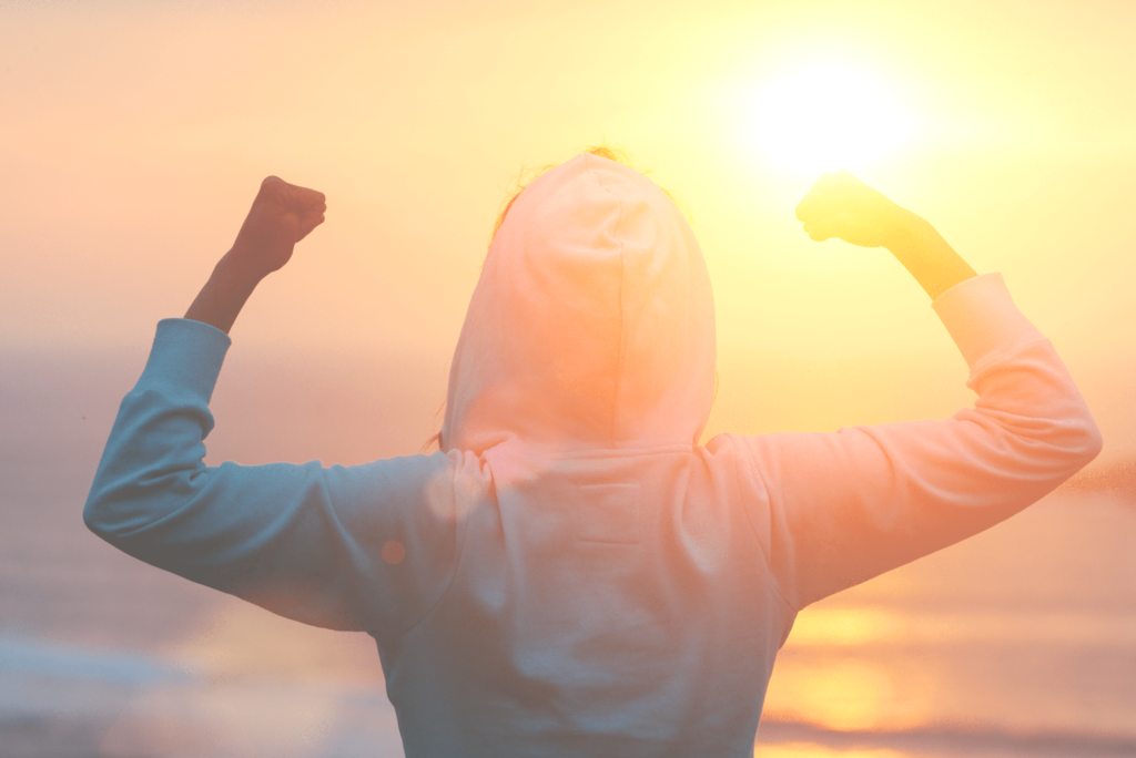 Man or woman in hoodie standing on the beach looking at the sea and raising their hands in the air with motivation