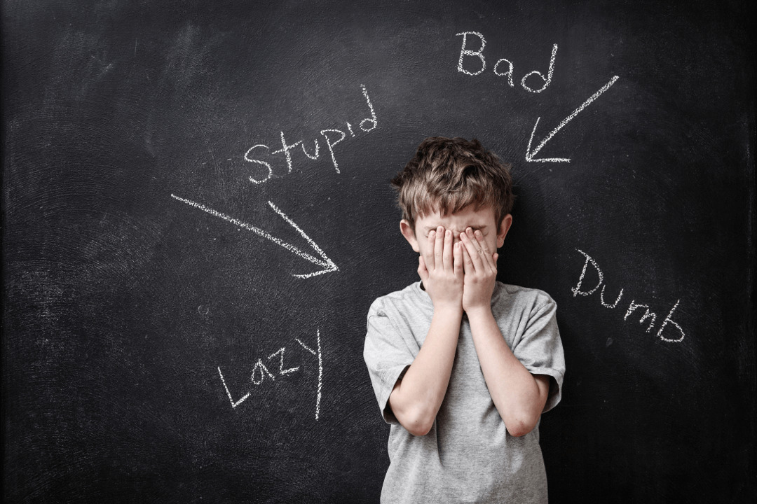 boy stands with his hands in front of his face in front of a blackboard on which there are catchwords pointing at him.