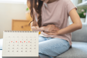 Frau sitzt mit schmerzendem Bauch auf dem Bett. Im Vordergrund steht ein Kalendar, in dem die Tage der Periode gekennzeichnet sind.