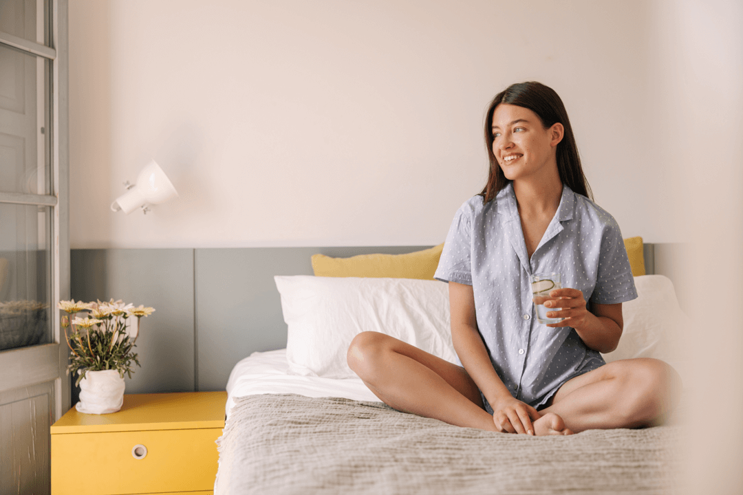 Woman sitting smiling on bed with glass in hand. Without adenomyosis pain.