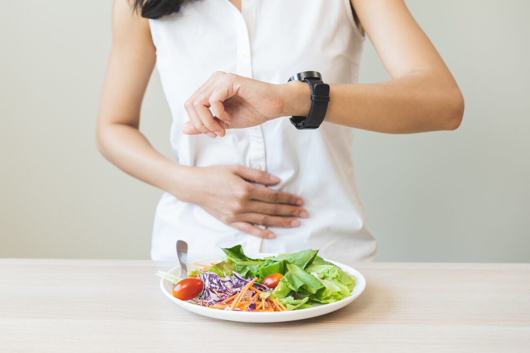 Frau steht vor einem Tisch, auf dem ein Teller voll mit Gemüse/Lebensmitteln steht und schaut auf die Uhr. Sie hält sich den Bauch vor Hunger