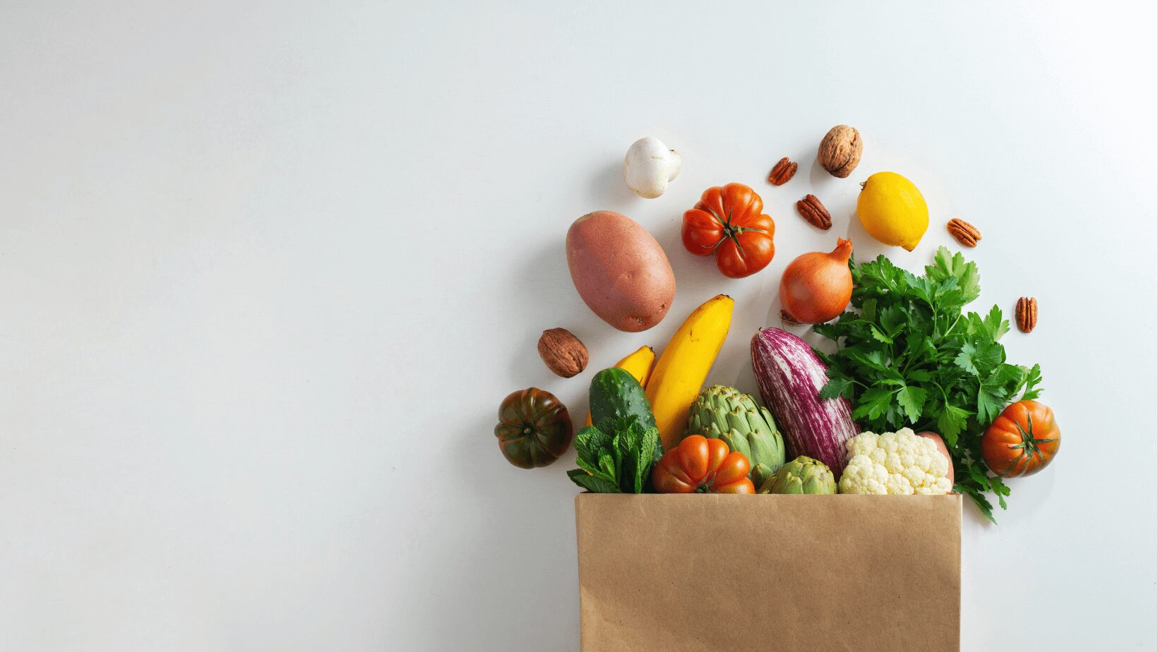 brown shopping bag on a white background with lots of fruit and vegetables falling out of it
