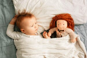 Baby is lying in bed with a doll next to him/her. Both have small red spots on their faces, showing the chickenpox rash.